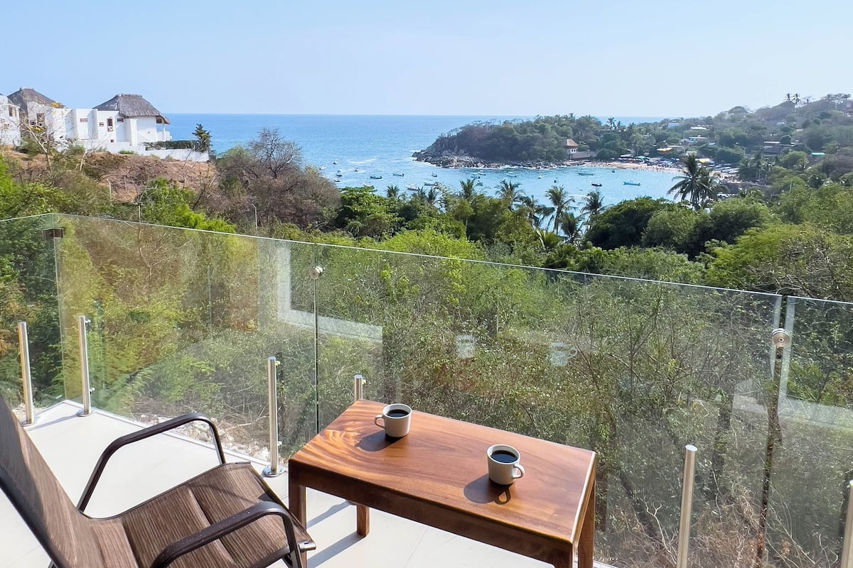Ein Balkon mit Tisch und Stühlen, der Blick auf die Küste mit Booten und Strand bietet.