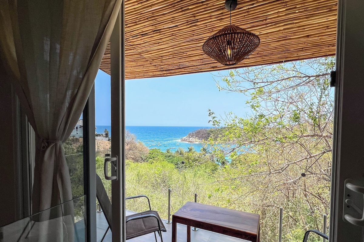 Ein Balkon mit Tisch und Stühlen, der auf eine Küstenlandschaft mit Bäumen und dem Meer blickt.