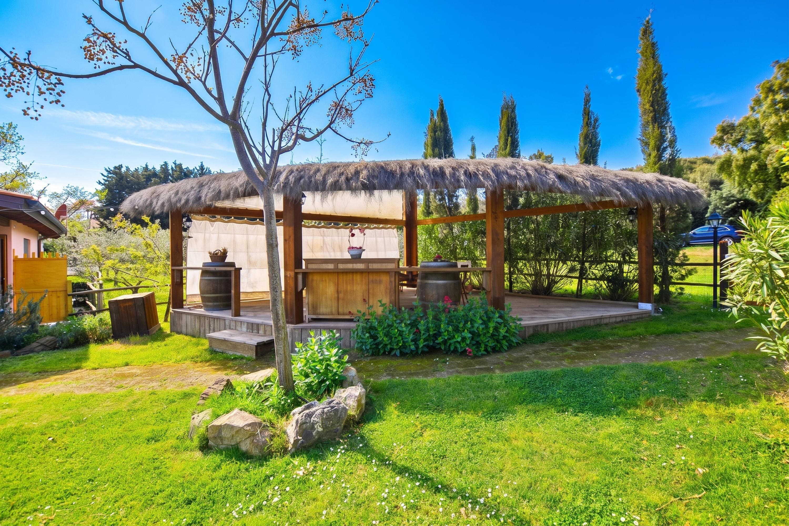 A wooden gazebo with a thatched roof, surrounded by a well-maintained garden with grass, rocks, and flowering plants.