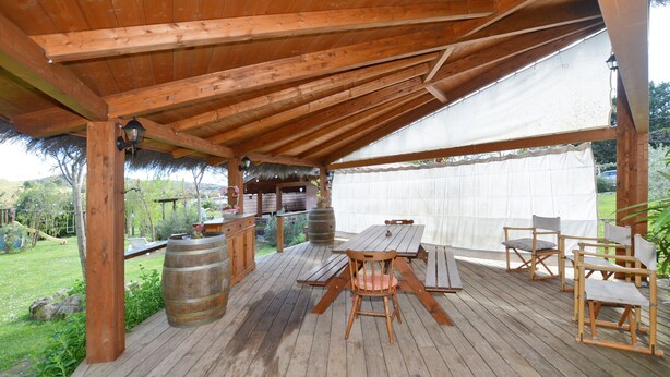 Eine Holzterrasse mit Tisch, Stühlen, einem Fass und einem strohgedeckten Bauwerk.