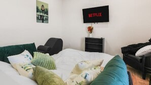 Un dormitorio con una cama, una silla, un televisor montado en la pared y una mesita de noche con un florero.