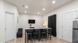 Habitación pequeña con kitchenette, zona de comedor con cuatro sillas, televisor montado en la pared y una puerta a la izquierda.