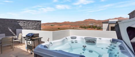 A hot tub with a view of a desert landscape, a stone wall, and a grill with chairs.