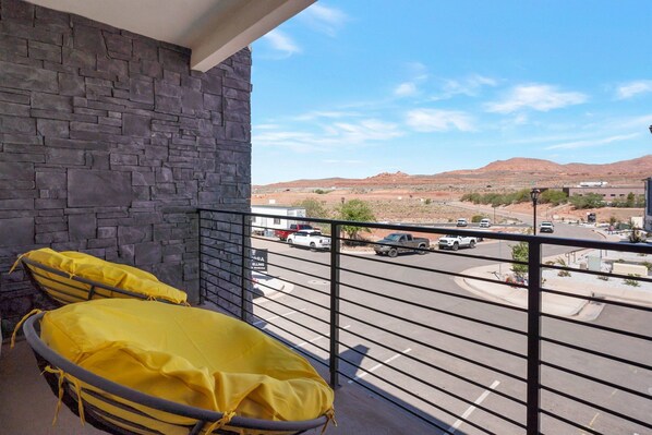 A balcony with a stone wall and two yellow-covered chairs. A street with cars and a distant mountain range is visible.