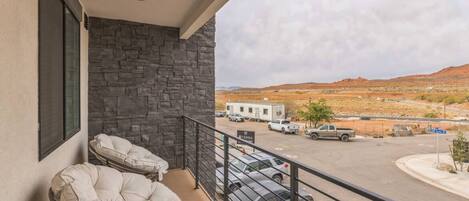 A balcony with two cushioned chairs, a stone wall, and a view of a parking lot and distant hills.