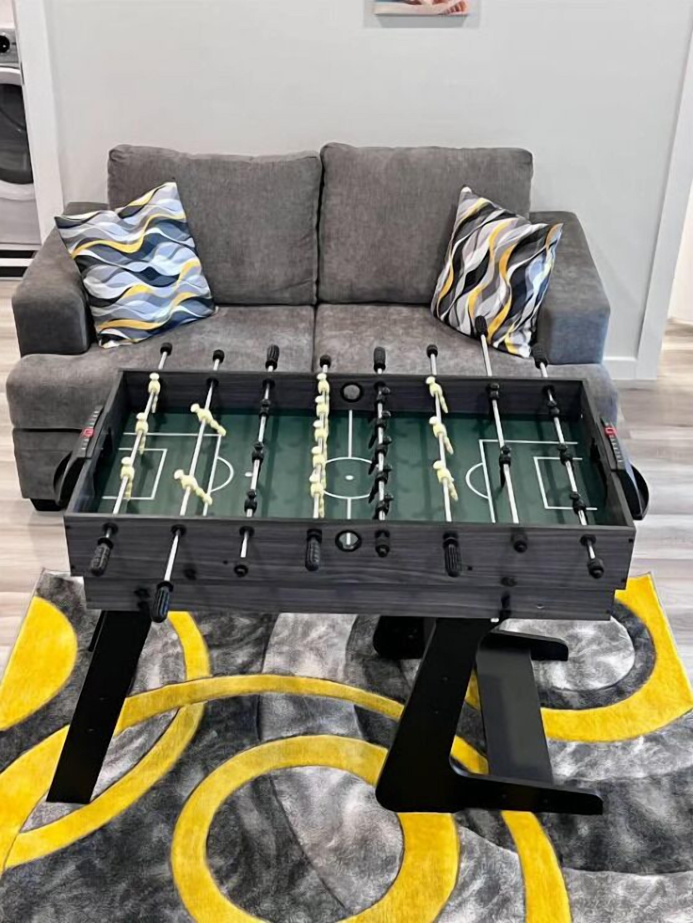 A foosball table in a living room with a gray sofa and patterned cushions.