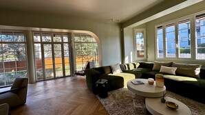 A spacious living room with a large sectional sofa, a round coffee table, and a dining table. There are large windows providing natural light and views of the outside. - Dream villa 50 meters from the beach! (Benerville-sur-Mer)