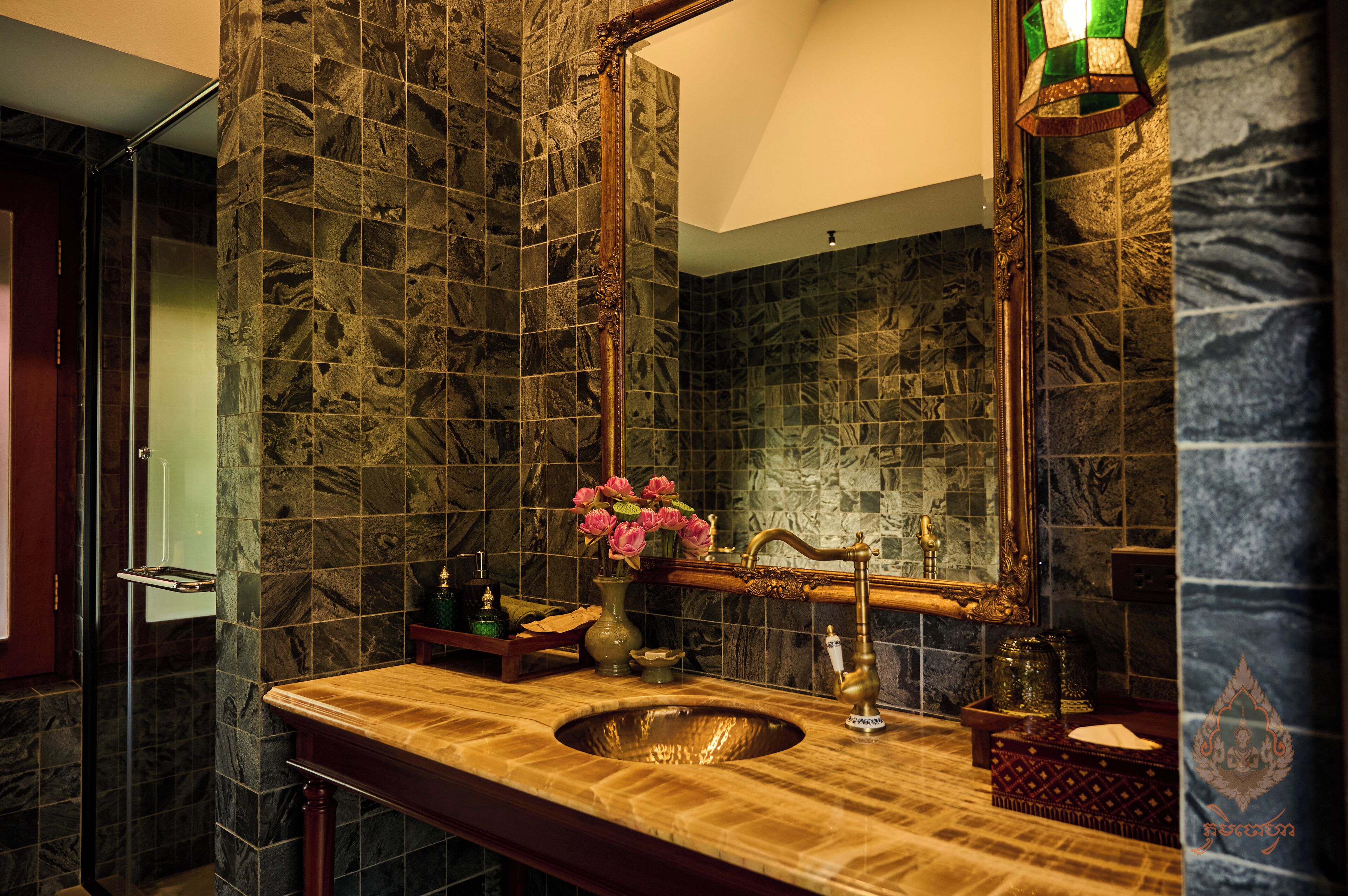 Une salle de bain avec un mur recouvert de carreaux de pierre, un meuble-lavabo en bois, un lavabo, un miroir et un vase avec des fleurs.