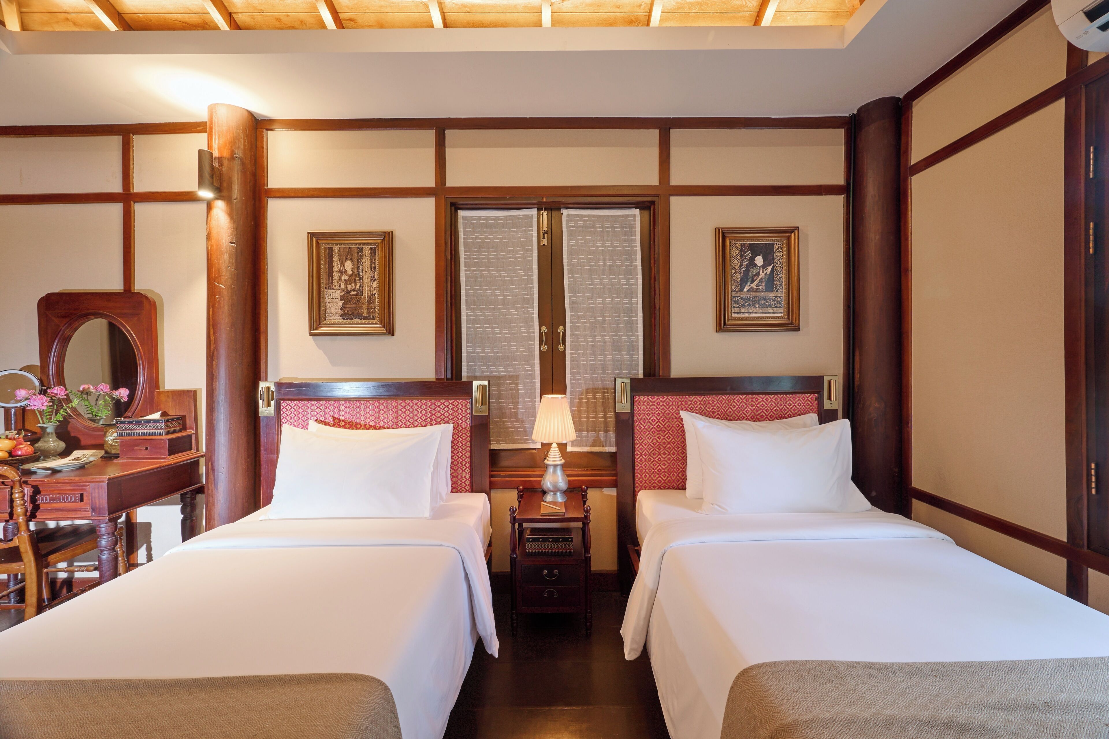 Quarto de hotel com duas camas, uma escrivaninha de madeira, um espelho e duas imagens emolduradas nas paredes.
