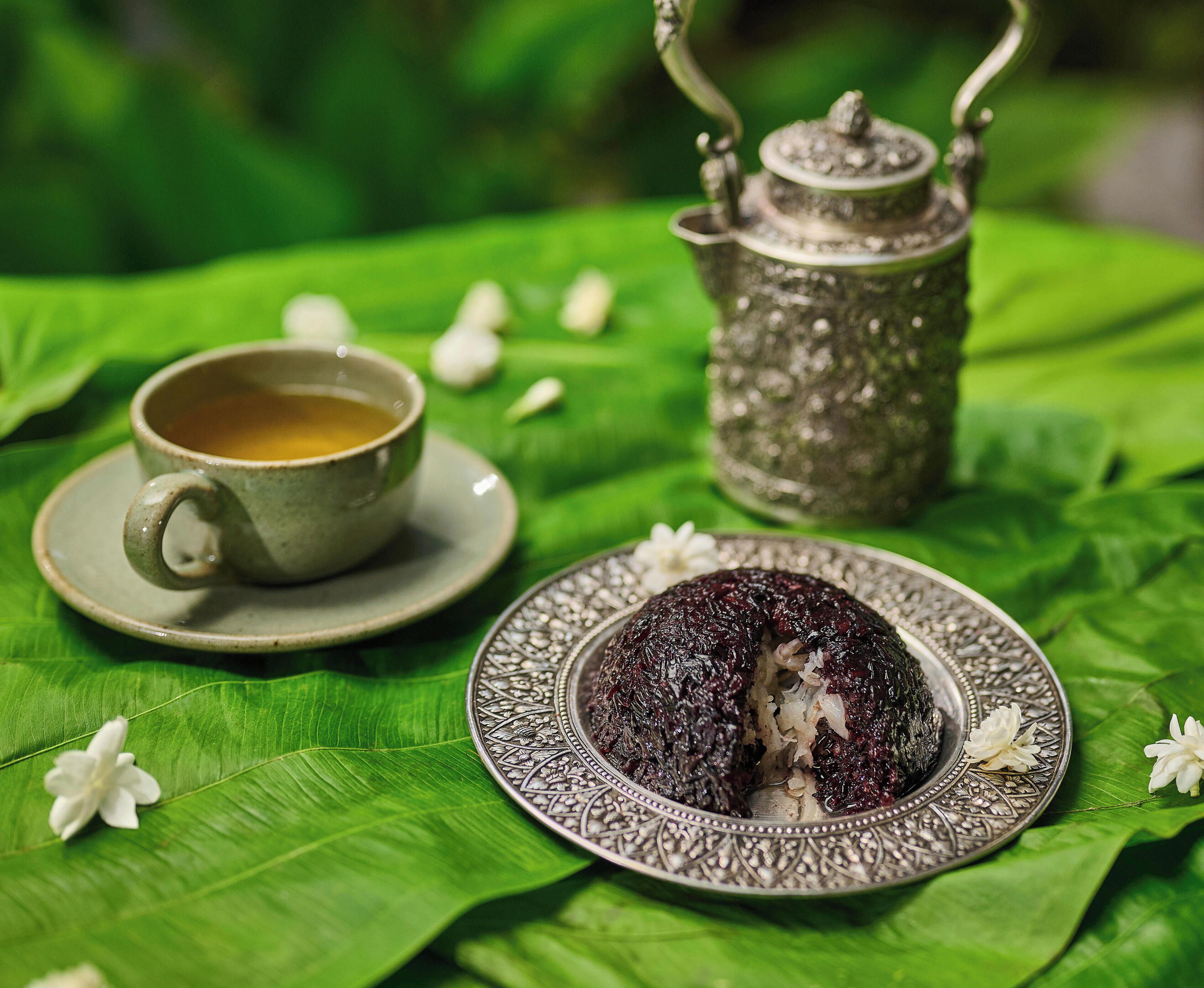 A cup of tea, a teapot, and a plate with food on a green leaf.