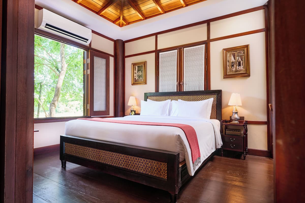 A bedroom with a large bed, wooden paneling, a nightstand with a lamp, and a window with a view of trees.