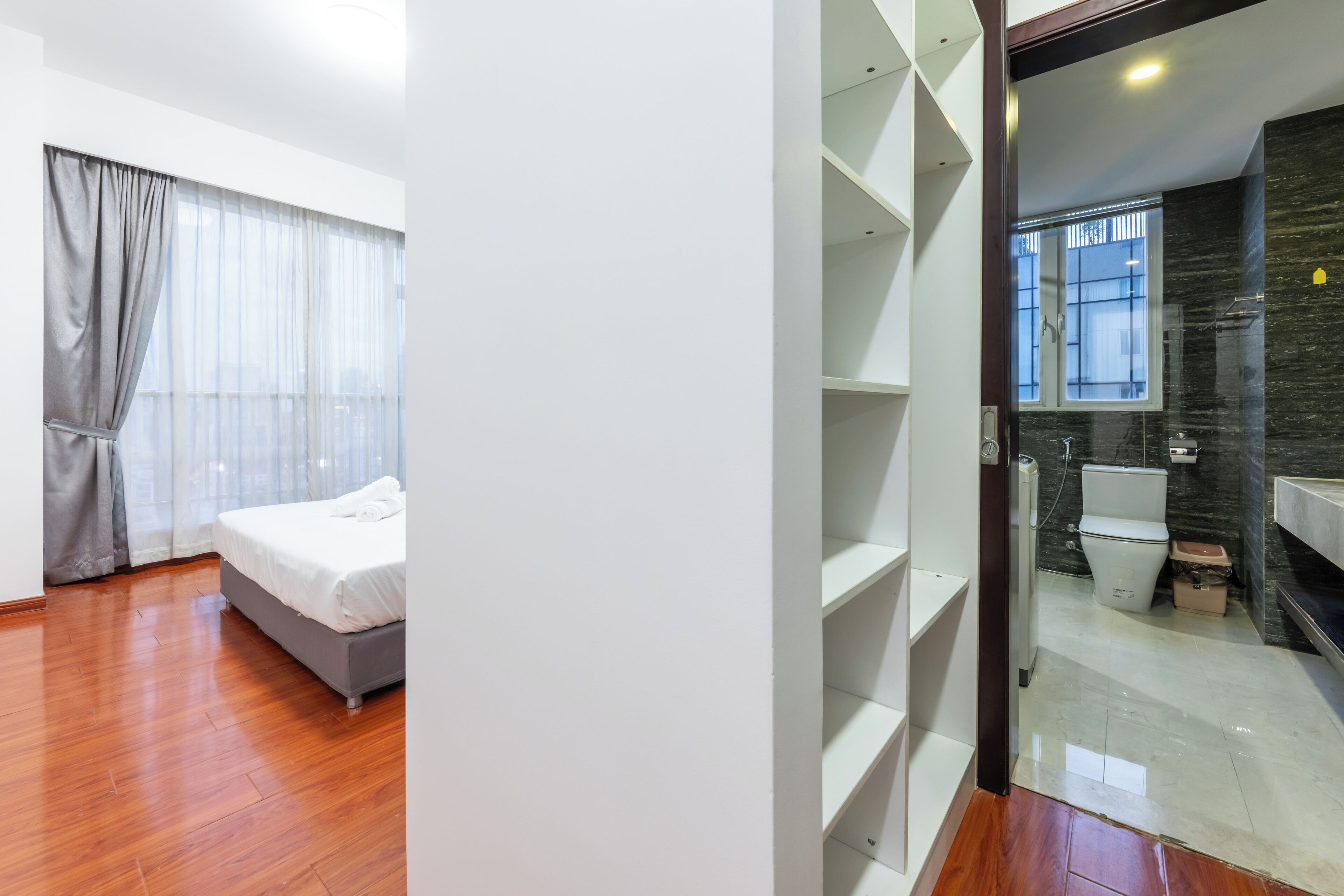Una habitación de hotel moderna con piso de madera, una cama, un ventanal con cortinas y un baño con inodoro y lavamanos.
