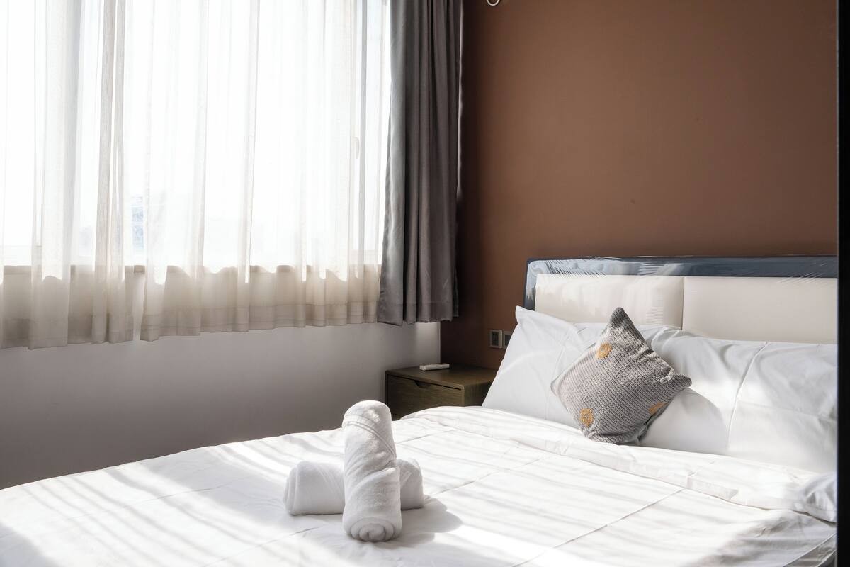 Una cama bien hecha con sábanas blancas y una toalla enrollada, una mesita de noche de madera con un control remoto, y una ventana con cortinas traslúcidas.