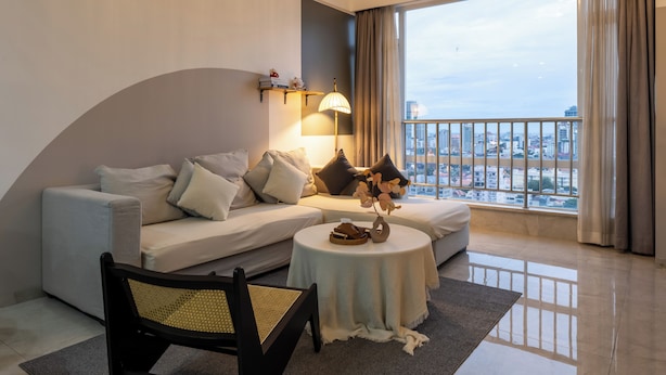 Une chambre d’hôtel moderne, dotée d’un canapé, d’une table ronde, d’une chaise, d’une lampe et offrant une vue sur le paysage urbain grâce à de grandes fenêtres.