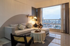 A modern hotel room with a sofa, a round table, a chair, a lamp, and a view of the cityscape through large windows. - Yue Tai Apartment City Center by Soben (Phnom Penh)