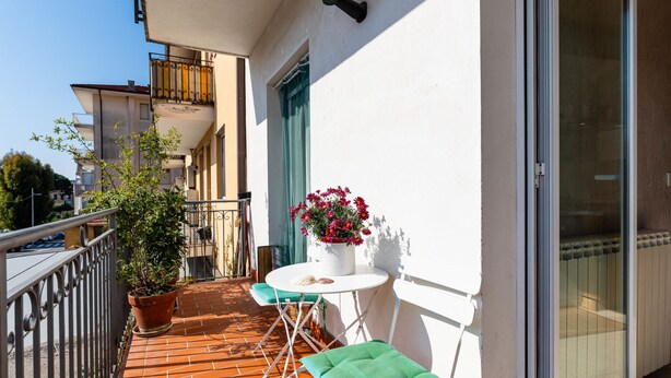 A balcony with a tiled floor, a potted plant, a table with a chair, and a window with a curtain.