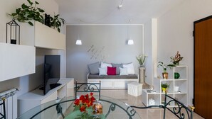 A modern living room with a glass table, a sofa with cushions, a TV, and a bookshelf.