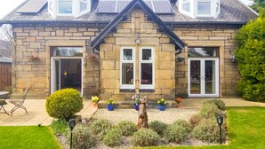 A stone house with a gabled roof, solar panels, and a well-maintained garden with potted plants and a wooden sculpture.