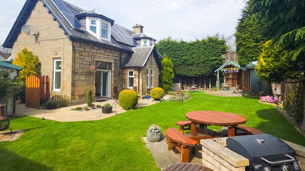 A stone house with a sloped roof, a patio with a barbecue, and a well-maintained lawn.