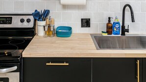 A modern kitchen with white tiles, wooden shelves, a stainless steel sink, and a black stove. - Cozy Retro Gem for the Fam W/ AC (Near Roger&WEM) (Edmonton)