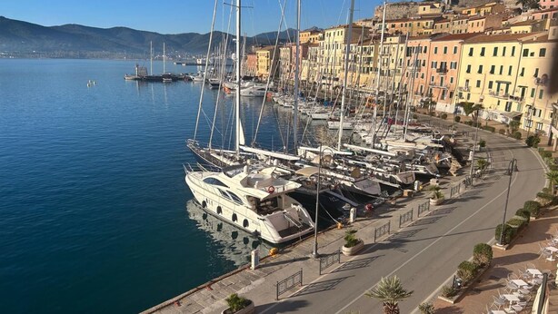 Un port de plaisance où sont amarrés de nombreux voiliers, une promenade en bord de mer avec des terrasses, et des bâtiments aux couleurs vives en arrière-plan.