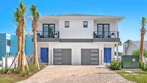 Una casa moderna de dos pisos con garaje, un balcón con sillas y palmeras.