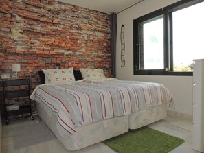 A bedroom with a brick wall, a bed with striped bedding, a nightstand with a lamp, and a window with a view.