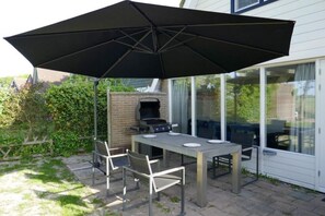 Une terrasse extérieure avec une table, des chaises, un barbecue et un grand parasol.