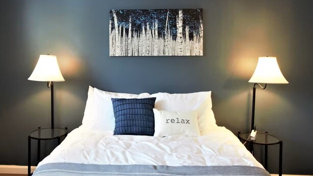 A neatly made bed with white bedding and a blue pillow, two bedside lamps, and a painting of a forest on the wall. - Oak Overflight Oasis Condo-Minutes to WPAFB & WSU with AC, fast WiFi (Fairborn)