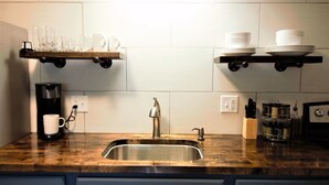 A kitchen with a stainless steel sink, wooden countertop, and two floating shelves holding dishes and glasses. - Oak Overflight Oasis Condo-Minutes to WPAFB & WSU with AC, fast WiFi (Fairborn)