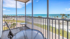 A balcony with a glass table and chairs, overlooking a beach with palm trees and a clear blue sky. - GULF VIEW | 2 QUEEN BEDS | SLEEPS 4 | 5TH FLOOR BALCONY | STEPS TO BONITA BEACH (Bonita Springs)