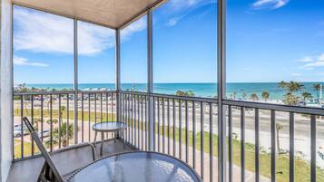 A balcony with a glass table and chairs, overlooking a beach with palm trees and a clear blue sky.