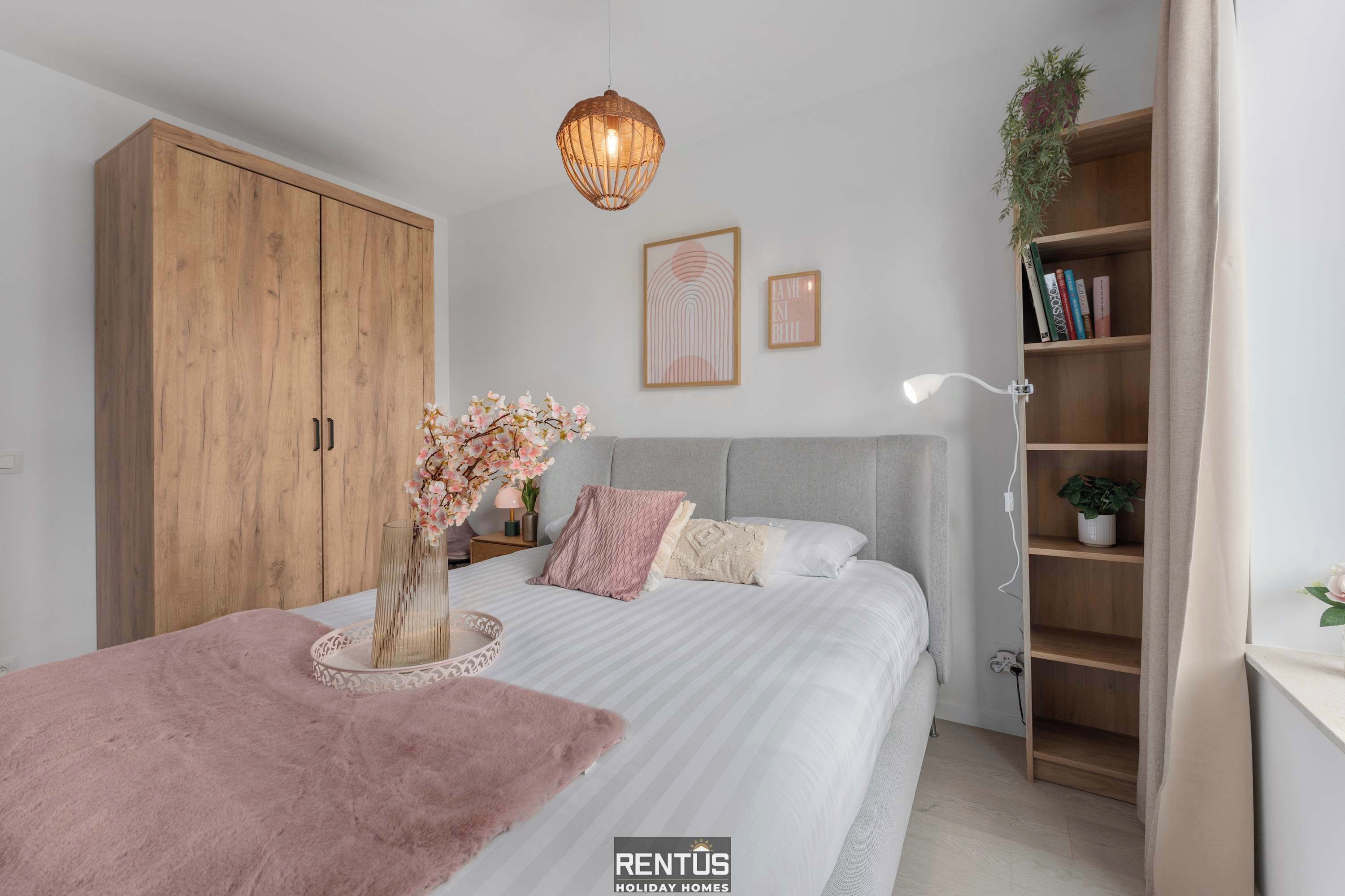 A bedroom with a bed, a wardrobe, a bedside table with a lamp, a bookshelf, and a hanging light fixture.