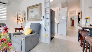 Een moderne woonkamer met een grijze bank, een tafeltje met een lamp en een boekenkast. Achtergrond toont een keuken met een koelkast en een eethoek met bankjes.