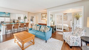 A spacious living area with a blue sofa, wooden coffee table, and patterned armchair. A kitchen with white cabinets and a dining area with bar stools is visible in the background. - 3 Mi to Duck Lake State Park: Home w/ Deck! (Muskegon)