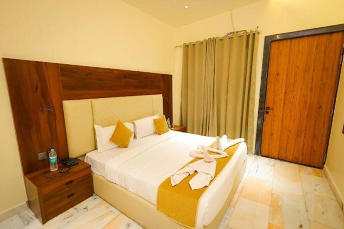 Habitación de hotel con cabecera de madera, cama con ropa de cama blanca y detalles amarillos, mesita de noche con una botella y un teléfono, y puerta con marco de madera.