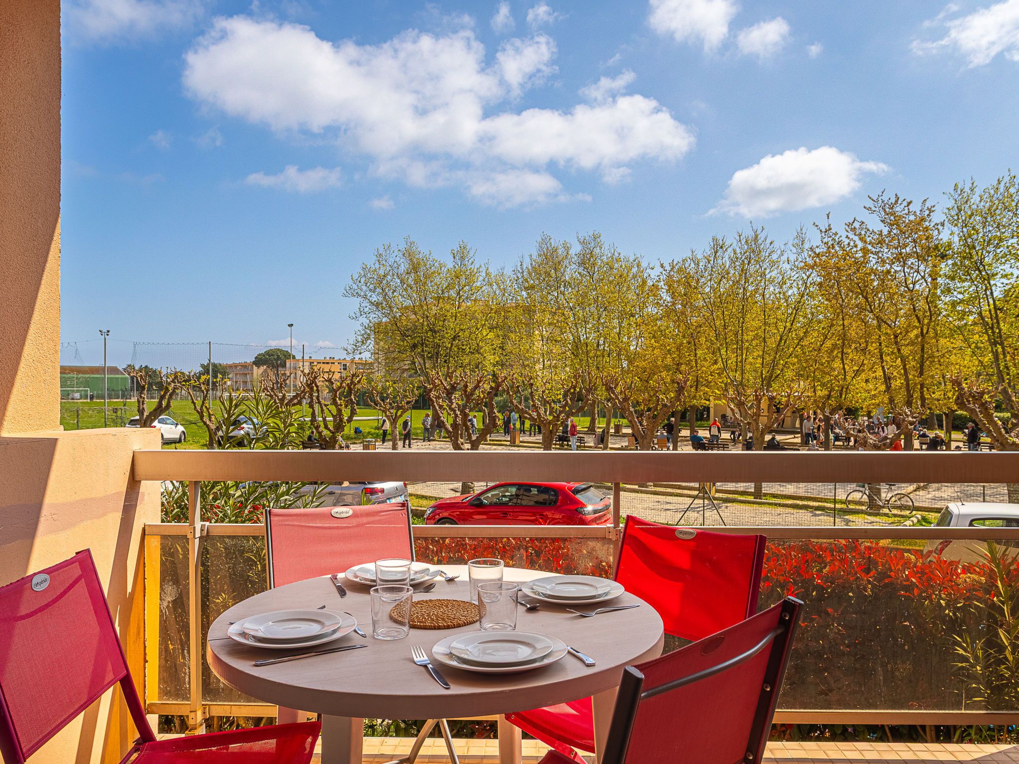 Un balcón con una mesa lista para comer, con vista a un parque con árboles y una calle.
