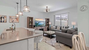 A modern living room with a grey sofa, a TV, and a kitchen area with a bar. - Magical Themed Home with Pool in Top Resort (Davenport)