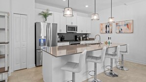 A modern kitchen with white cabinets, stainless steel appliances, a central island with bar stools, pendant lighting, and artwork on the wall. - Magical Themed Home with Pool in Top Resort (Davenport)