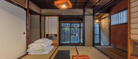 Ein traditioneller japanischer Raum mit Tatami-Bodenbelag, einem Futon-Bett, einem schwarzen Tisch und einer Schiebetür, die zu einem Garten führt.