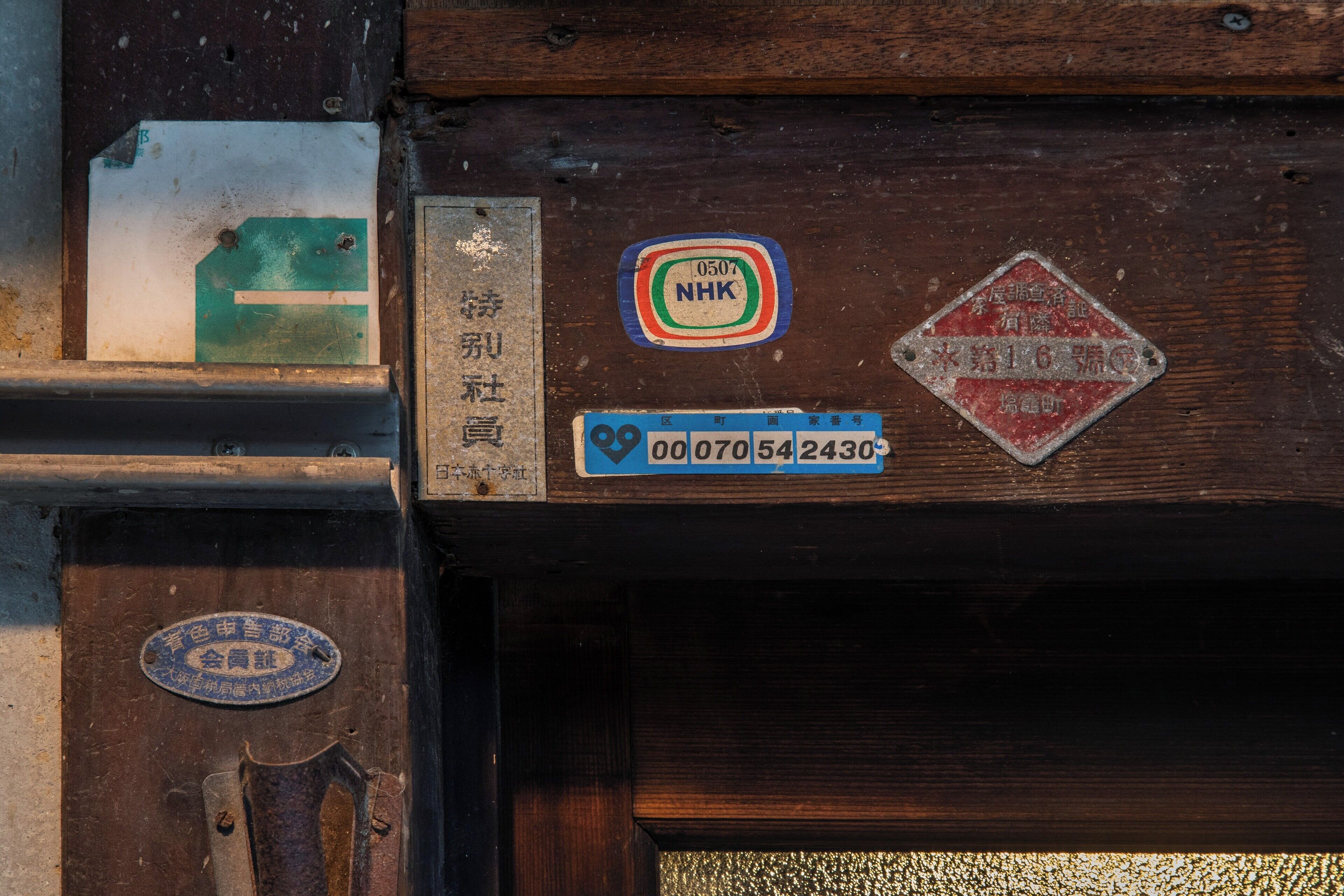 A wooden door with various signs and labels, including a blue and white NHK logo, a red diamond-shaped sign, and a blue rectangular sign with numbers.