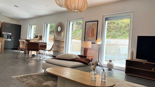 Una sala de estar moderna con un sofá, una mesa de centro y un televisor. Se observan amplias ventanas, un comedor y una cocina al fondo.