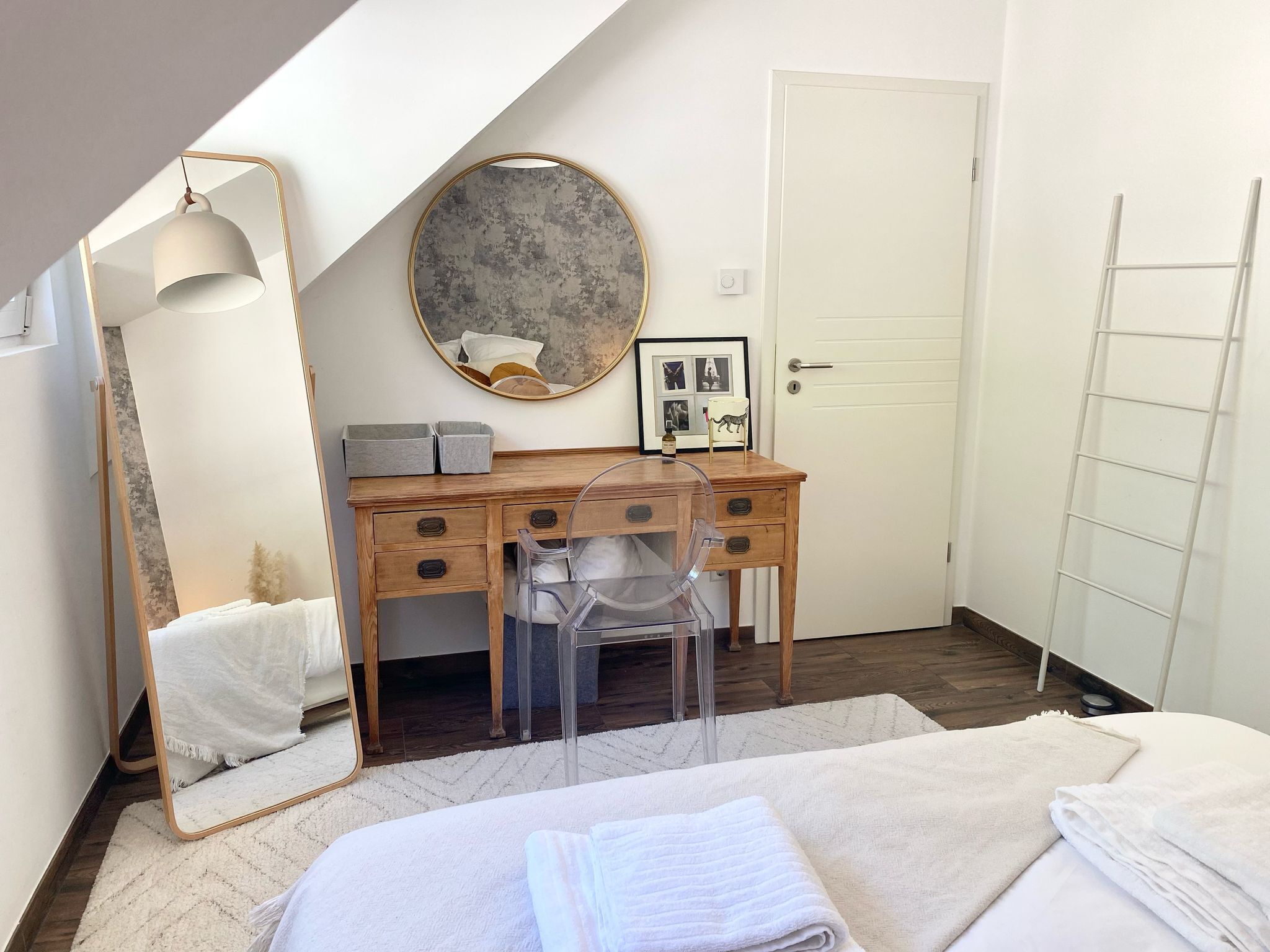 Une chambre à coucher comprenant un lit, un bureau avec une chaise, un grand miroir, une horloge ronde fixée au mur et une porte.