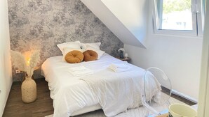 Un dormitorio con techo inclinado, una cama con ropa blanca y varios almohadones, una mesita de noche con lámpara y una ventana con cortinas.
