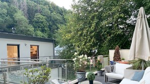 Un patio al aire libre moderno con barandilla de vidrio, mobiliario blanco y plantas en macetas.