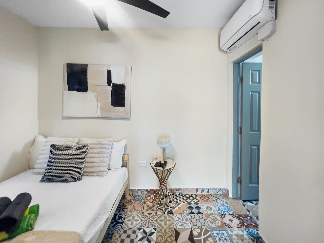A bedroom with a bed, a small side table with a lamp, a wall-mounted air conditioner, and a patterned floor.