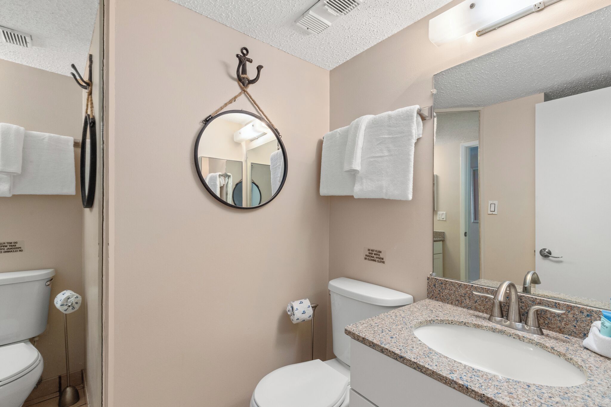 A bathroom with a toilet, sink, and mirror. There are towels hanging on hooks and a sign on the wall.