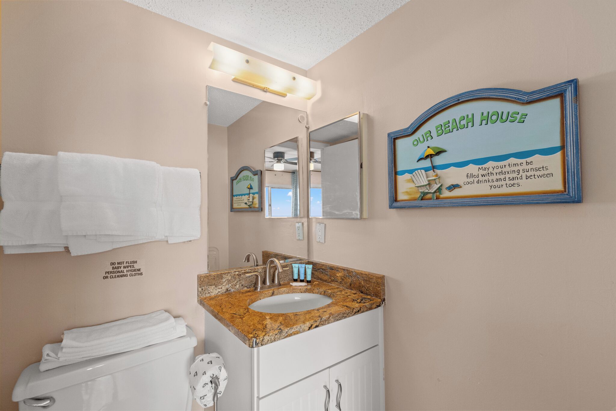 Une salle de bain équipée d’un lavabo, d’un miroir et d’un porte-serviettes. Un panneau est accroché au mur, sur lequel on peut lire : « Notre maison de plage ».