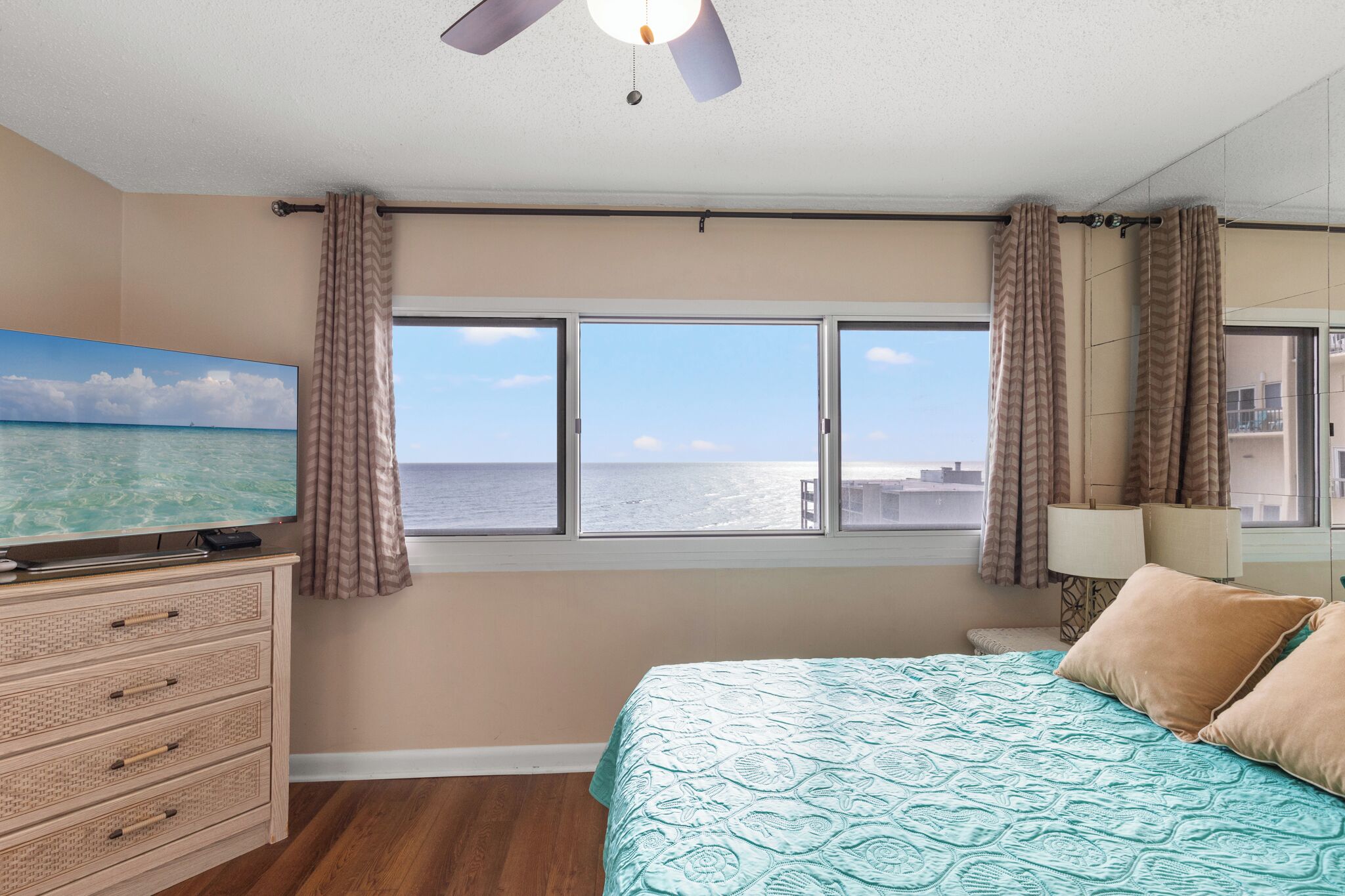 Un dormitorio con cama, televisor, ventilador de techo, ventana con cortinas y vista al mar.