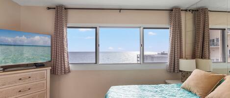 Un dormitorio con cama, televisor, ventilador de techo, ventana con cortinas y vista al mar.